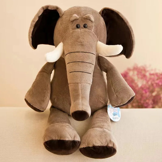 Um elefante de pelúcia marrom com orelhas grandes e presas brancas está sentado em um sofá claro, com flores cor-de-rosa desfocadas ao fundo.