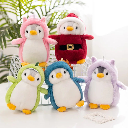 Cinco pelúcias de pinguins vestidos com fantasias coloridas, incluindo rosa, verde, azul, roxa e um de vermelho com a roupa de Papai Noel, em um ambiente com plantas.